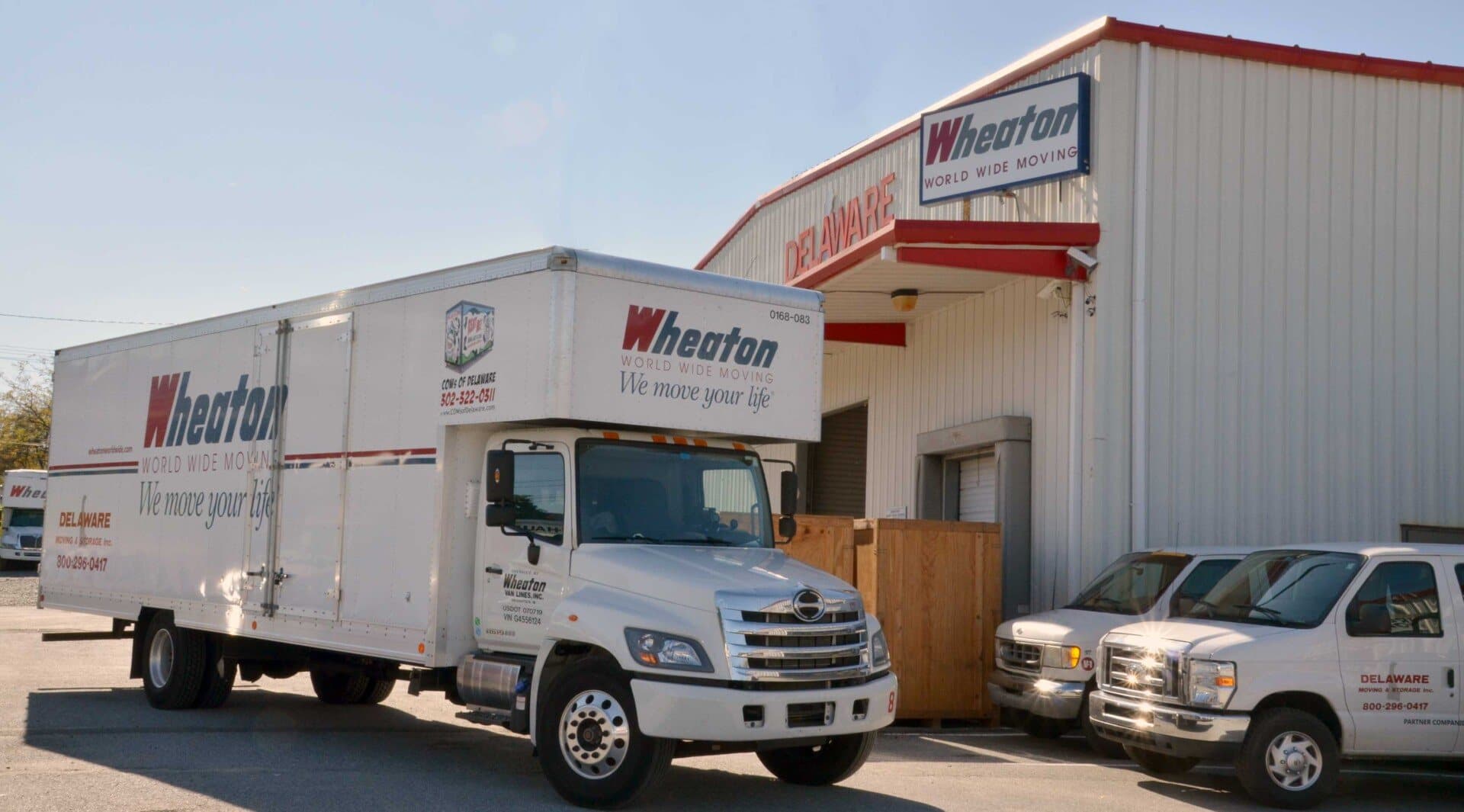 A branded Wheaton World Wide Moving truck and Delaware Moving and Storage vans parked outside the company facility in Delaware